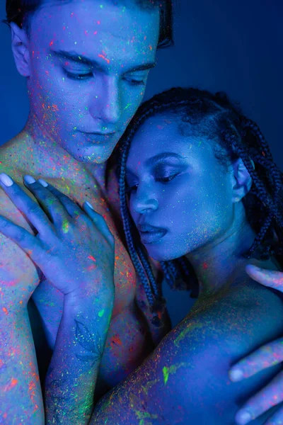 Sensuale coppia interrazziale in vernice colorata corpo che abbraccia su sfondo blu con illuminazione ciano, bello uomo a petto nudo e incantevole donna africana americana con dreadlocks — Foto stock