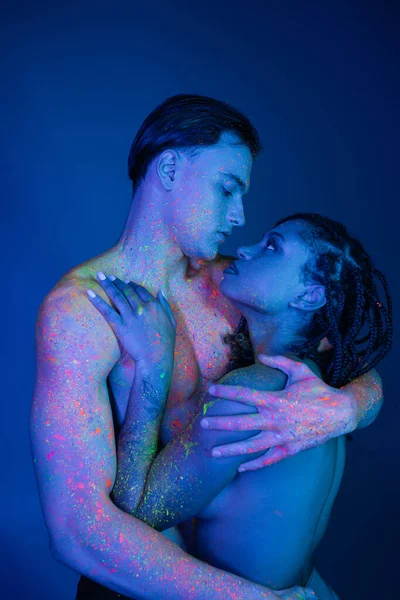 Youthful interracial couple in colorful neon body paint embracing on blue background with cyan lighting, shirtless man with muscular torso and african american woman with dreadlocks — Stock Photo