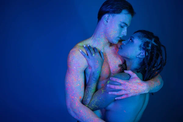 Multicultural couple in colorful neon body paint hugging and looking at each other, shirtless man with muscular torso and african american woman with dreadlocks on blue background with cyan lighting — Stock Photo