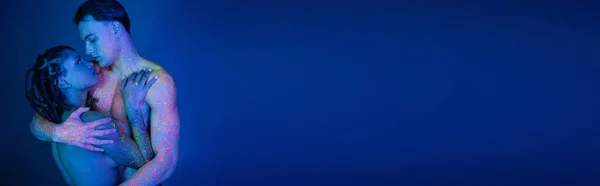 Pareja interracial joven y desnuda en colorida pintura corporal de neón abrazándose sobre fondo azul con iluminación cian, momento íntimo de encantadora mujer afroamericana y hombre con cuerpo muscular, pancarta - foto de stock