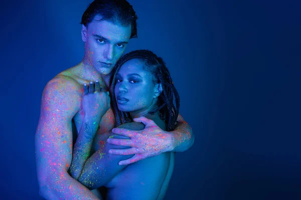 Multicultural couple in colorful neon body paint embracing and looking at camera on blue background with cyan lighting, shirtless man with muscular body and african american woman with dreadlocks — Stock Photo