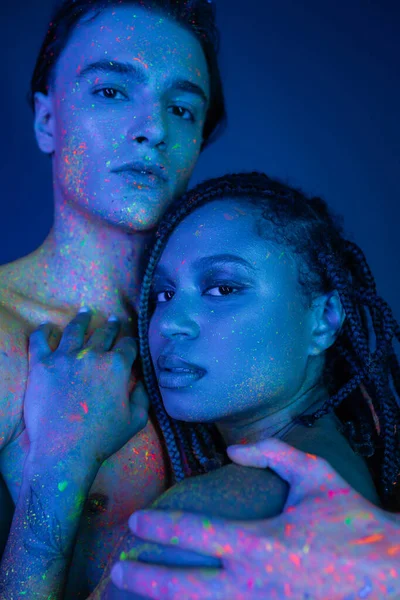 Youthful interracial couple in colorful neon body paint hugging and looking at camera on blue background with cyan lighting, confident shirtless man and african american woman with dreadlocks — Stock Photo
