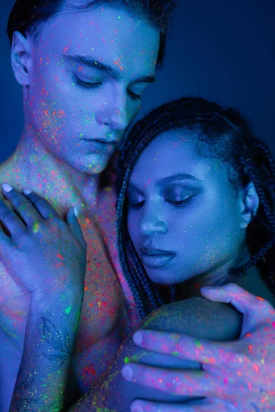 Young and sensual interracial couple in colorful body paint embracing on blue background with cyan lighting, bare-chested man and african american woman with dreadlocks and naked shoulders — Stock Photo
