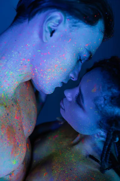 Momento íntimo de joven pareja multicultural en colorida pintura corporal de neón, hombre de pecho desnudo y sexy mujer afroamericana sobre fondo azul con iluminación cian - foto de stock