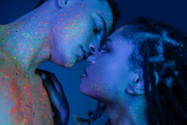 Low angle view of multicultural couple in colorful neon body paint standing face to face on blue background with cyan lighting, captivating african american woman and bare-chested man — Stock Photo