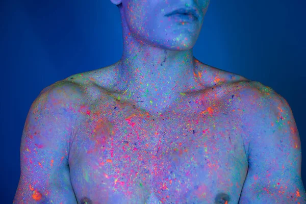 Partial view of shirtless, bare-chested man with muscular body posing in multicolored and bright neon body paint while standing on blue background with cyan lighting effect — Stock Photo