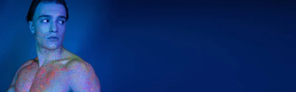 Jovem e atraente homem sem camisa em tinta de corpo de néon radiante e multicolorido olhando para longe, enquanto em pé no fundo azul com efeito de iluminação ciano, banner — Fotografia de Stock