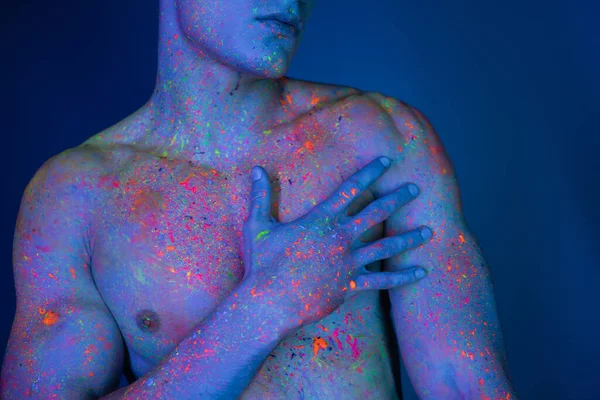 Vista recortada de un hombre joven, sin camisa y con el pecho desnudo tocando el cuerpo muscular mientras está de pie en pintura de cuerpo de neón colorido y magnético sobre fondo azul con efecto de iluminación cian - foto de stock