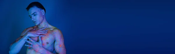 Jovem e carismático homem sem camisa em neon brilhante corpo pintura tocando corpo muscular enquanto posando em fundo azul com efeito de iluminação ciano, banner — Fotografia de Stock
