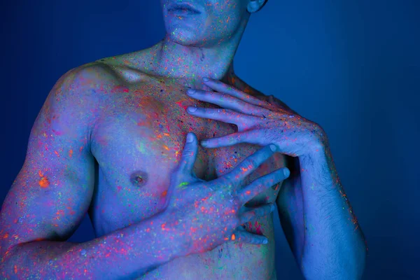 Visão parcial do homem sem camisa com tronco muscular tocando peito nu enquanto posando em tinta corporal de néon colorido e vibrante no fundo azul com efeito de iluminação ciano — Fotografia de Stock