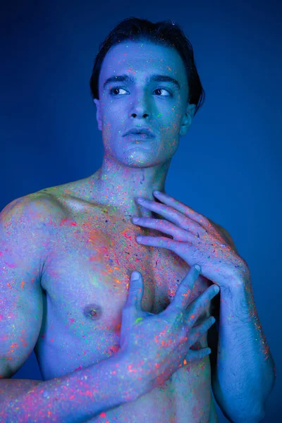 Jeune, homme torse nu et accrocheur posant dans la peinture au néon colorée vibrante, touchant poitrine nue et regardant loin sur fond bleu avec effet de lumière cyan — Photo de stock