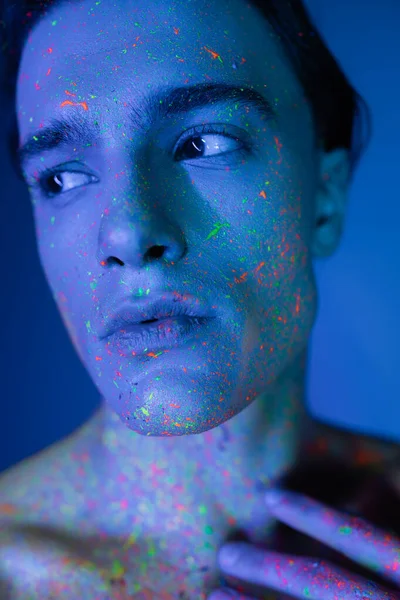 Primer plano retrato de un hombre joven y guapo en pintura corporal de neón radiante y multicolor mirando hacia otro lado sobre fondo azul con efecto de iluminación cian - foto de stock