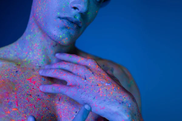 Vue recadrée de l'homme torse nu tenant les mains près de la poitrine nue tout en posant dans de la peinture au néon colorée et vibrante sur fond flou et bleu avec effet d'éclairage cyan — Photo de stock