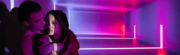Youthful and impassioned multicultural couple, sexy african american woman and youthful man embracing while standing on abstract purple background with neon rays and lighting effects, banner — Stock Photo