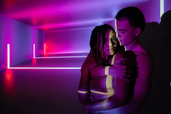 Young and confident man embracing captivating african american woman with dreadlocks on abstract purple background with neon rays and lighting effects — Stock Photo