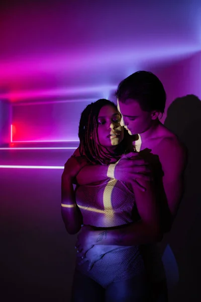 Hombre joven y guapo abrazando apasionada mujer afroamericana con rastas mientras está de pie sobre fondo púrpura abstracto con rayos de neón y efectos de iluminación - foto de stock