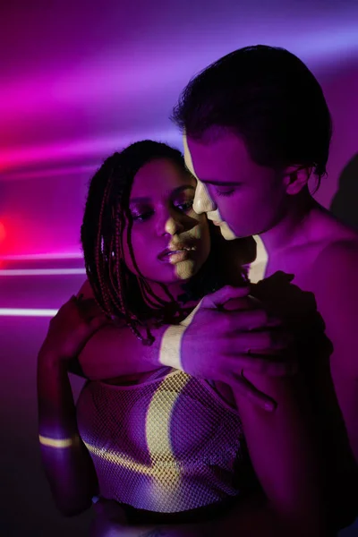 Jovem e apaixonado casal, mulher americana africana com dreadlocks e jovem homem bonito abraçando em fundo roxo abstrato com raios de néon e efeitos de iluminação — Fotografia de Stock