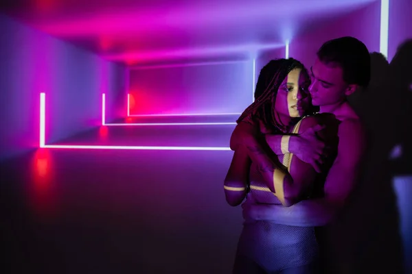 Impassioned interracial couple, charismatic man and youthful african american woman with dreadlocks embracing on abstract purple background with neon rays and lighting effects — Stock Photo