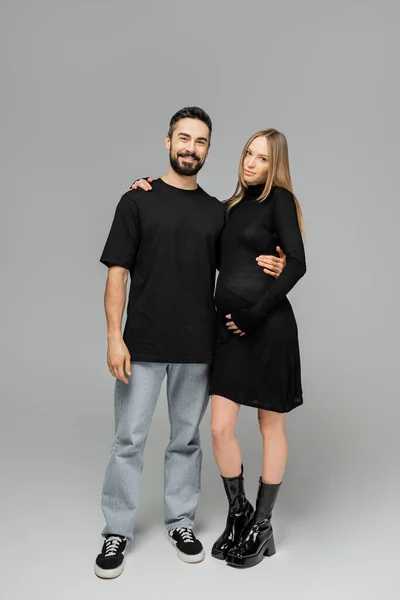 Volle Länge von fröhlichen bärtigen Mann in schwarzem T-Shirt und Jeans umarmt schwangere und stilvolle Frau in Kleid und stehen zusammen auf grauem Hintergrund, wachsende neue Lebenskonzept — Stockfoto