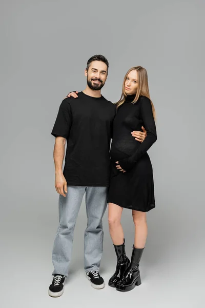 Pleine longueur d'homme barbu gai en t-shirt noir et jeans étreignant femme enceinte et élégante en robe et debout ensemble sur fond gris, concept de vie de plus en plus nouveau — Photo de stock