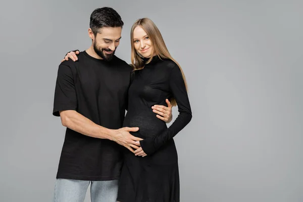 Позитивный бородатый мужчина в черной футболке касается живота светловолосой беременной жены в стильном платье и обнимает друг друга, стоя в одиночестве на серой, растущей новой жизненной концепции — стоковое фото