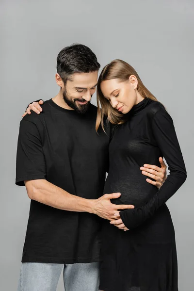 Улыбающийся мужчина в черной футболке и джинсах, касающийся живота беременной и стильной жены, стоя и обнимая друг друга изолированно на серой, растущей новой жизненной концепции — стоковое фото