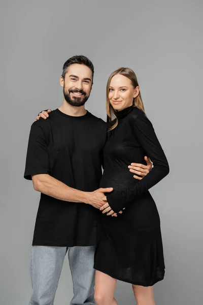 Положительная беременная женщина в стильном черном платье, смотрящая в камеру и обнимая бородатого мужа в футболке и джинсах, стоящего изолированно на серой, растущей новой концепции жизни — стоковое фото