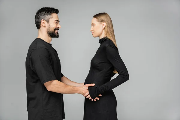 Вид сбоку на позитивного бородатого мужчину в футболке, касающегося живота беременной жены в стильном черном платье и смотрящего друг на друга, стоя изолированно на сером, растущем новом жизненном концепте — стоковое фото