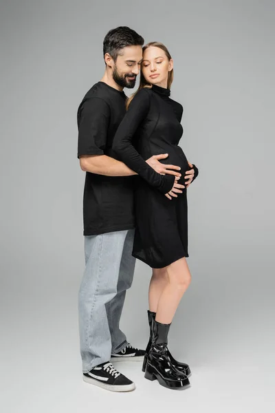 Comprimento total do homem barbudo sorridente em jeans e camiseta abraçando a esposa elegante e grávida em vestido preto enquanto estão juntos em fundo cinza, crescendo novo conceito de vida — Fotografia de Stock