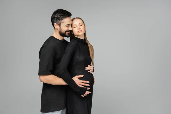 Радостный бородатый мужчина в футболке, обнимающий живот стильной и расслабленной беременной жены в черном платье, стоя с закрытыми глазами, изолированными от серого, растущей новой жизненной концепции — стоковое фото