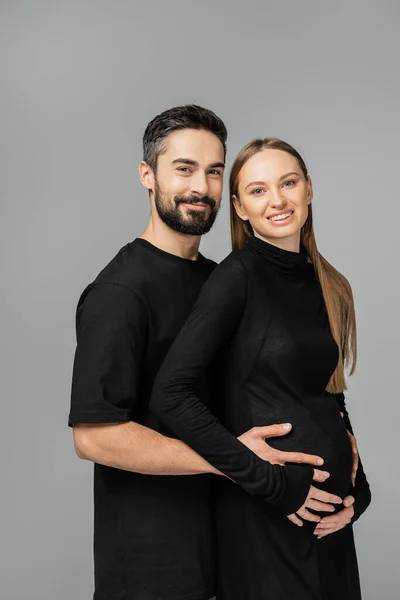 Retrato de hombre barbudo en camiseta negra mirando a la cámara mientras toca el vientre de la esposa embarazada sonriente y elegante en vestido aislado en gris, creciendo nuevo concepto de vida - foto de stock