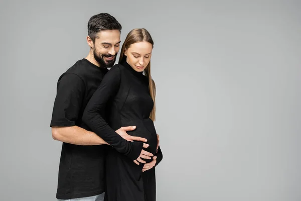 Positive bearded husband in t-shirt hugging belly of stylish and pregnant wife in black dress while standing together isolated on grey with copy space, growing new life concept — Stock Photo