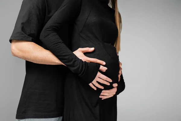 Обрезанный вид заботливого мужчины в черной футболке, обнимающего живот стильной беременной жены в платье и стоящего вместе изолированного на серой, растущей новой жизненной концепции — стоковое фото