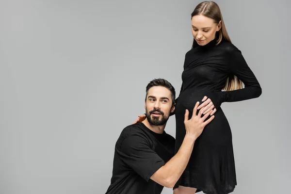 Захоплений бородатий чоловік у футболці, який торкається і слухає звуки живота стильної вагітної дружини в чорному платті, стоячи разом ізольовано на сірому, зростаючому новому життєвому концепті — стокове фото