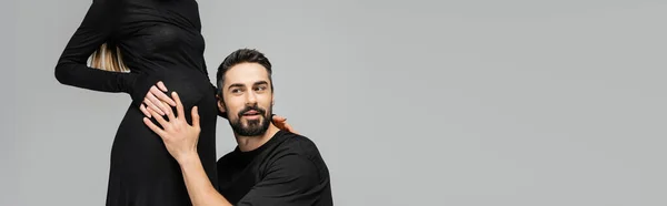 Excited and bearded husband in t-shirt listening and touching belly of pregnant and stylish wife in black dress isolated on grey with copy space, growing new life concept, banner — Stock Photo