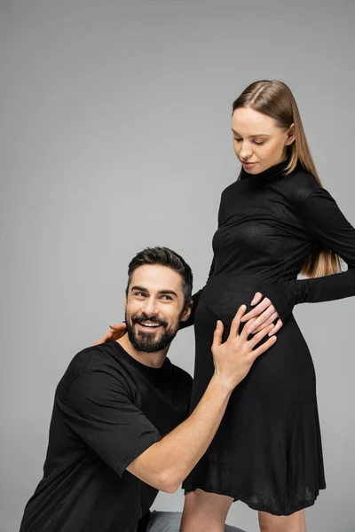 Homme barbu souriant en t-shirt écoutant le ventre de la femme à la mode et enceinte avec un maquillage naturel en robe noire élégante tout en restant isolé sur gris, un nouveau concept de vie en pleine croissance — Photo de stock