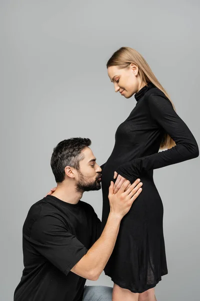 Seitenansicht eines bärtigen Mannes im schwarzen T-Shirt, der den Bauch einer stilvollen und lächelnden Schwangeren küsst, während er isoliert auf einem grauen, wachsenden neuen Lebenskonzept steht, Mann und Frau — Stockfoto