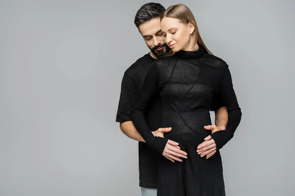 Бородатий чоловік в чорній футболці обіймає і зворушує живіт стильної і справедливої вагітної дружини в одязі, стоячи разом ізольовано на сірому, концепція народження дитини — стокове фото