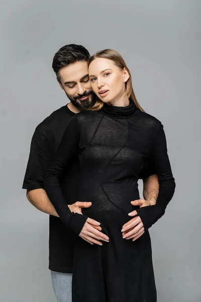 Bearded man in black t-shirt touching belly of stylish and pregnant fair haired wife in dress and standing together isolated on grey, concept of pregnancy and birth of child — Stock Photo