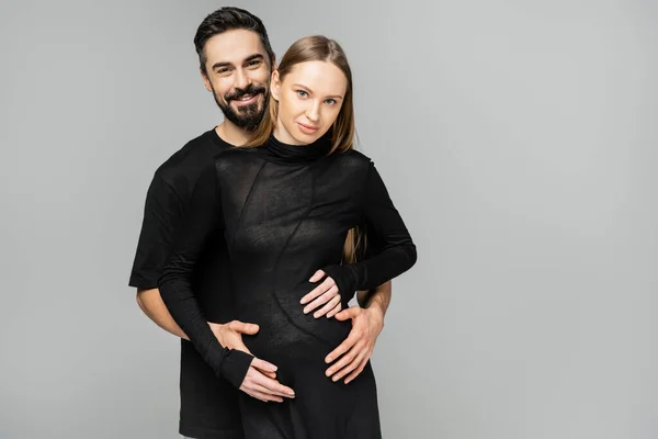 Позитивный и бородатый мужчина в черной футболке обнимает модную и беременную жену в черном платье и смотрит в камеру, стоя изолированно на сером, концепция рождения ребенка — стоковое фото