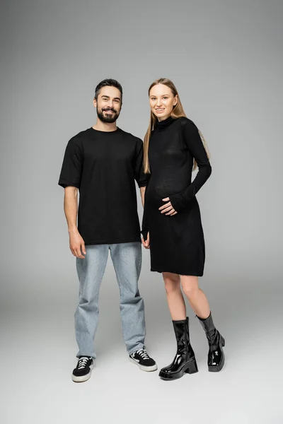 Полная длина веселой и модной беременной женщины в черном платье, смотрящей в камеру рядом с бородатым мужем в футболке и джинсах на сером фоне, концепция рождения ребенка — стоковое фото