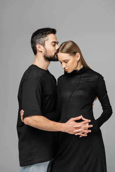 Бородатый муж в футболке, целующийся и трогательный живот стильной беременной жены в черном платье и одиноко стоящий на сером, концепция ожидающих родителей, счастливая семья — стоковое фото
