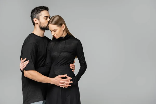 Бородатый мужчина в черной футболке целуется и держит за руку стильную беременную жену с закрытыми глазами и стоит изолированный на сером с копировальным пространством, концепция ожидающих родителей — стоковое фото