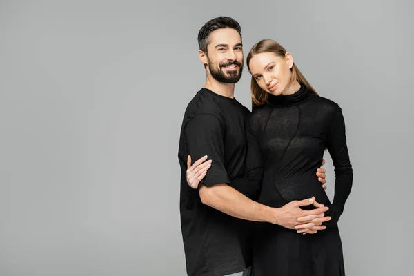 Positive and bearded husband in t-shirt looking at camera while hugging stylish pregnant wife in black dress isolated on grey, new beginnings and parenting concept — Stock Photo