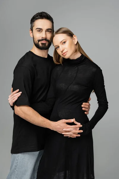 Mujer embarazada con estilo en vestido negro mirando a la cámara mientras toma de la mano del marido barbudo en camiseta aislada en gris, nuevos comienzos y el concepto de crianza - foto de stock