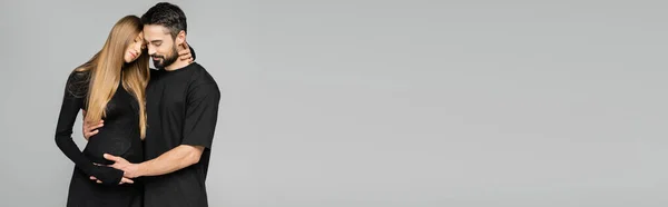 Marito barbuto in t-shirt nera che tocca la pancia della moglie alla moda dai capelli biondi e incinta mentre si trova isolato su grigio, nuovi inizi e concetto di genitorialità, banner — Foto stock