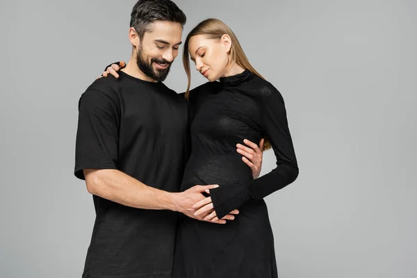 Радостный бородатый мужчина в черной футболке трогает живот модной беременной жены и смотрит вниз, стоя вместе изолированно на сером, новом начале и концепции воспитания, единения — стоковое фото