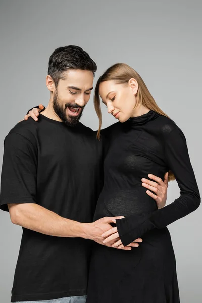 Homme barbu excité en t-shirt toucher le ventre et regarder femme enceinte à la mode en robe tout en se tenant ensemble isolé sur gris, nouveaux départs et concept parental — Photo de stock
