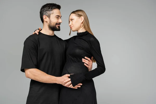 Улыбающийся и бородатый муж в черной футболке, обнимающий и смотрящий на стильную беременную жену в платье, стоя вместе изолированно на сером, новом начале и концепции воспитания — стоковое фото