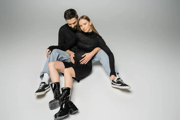 Homem barbudo em jeans e camiseta preta abraçando a mulher grávida na moda em vestido e botas enquanto se sentam juntos em fundo cinza, novos começos e conceito de parentalidade — Fotografia de Stock